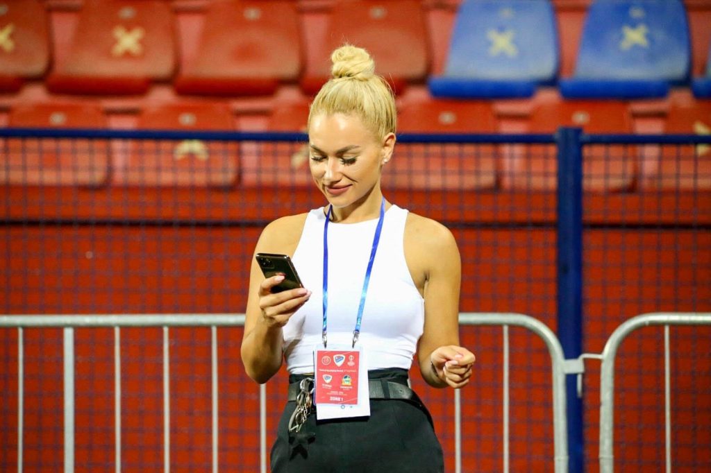 Žena plave kose u bijeloj majici stoji na stadionu i gleda u svoj mobilni telefon, noseći akreditaciju oko vrata. U pozadini se vide crvena i plava sjedišta na tribinama.